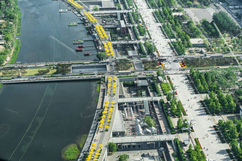 An aerial view of a riverside promenade with yellow trees, a bridge, and a large open plaza.