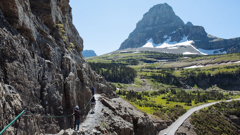 A mountain landscape with a prominent rocky cliff, a winding road, and a distant mountain peak.