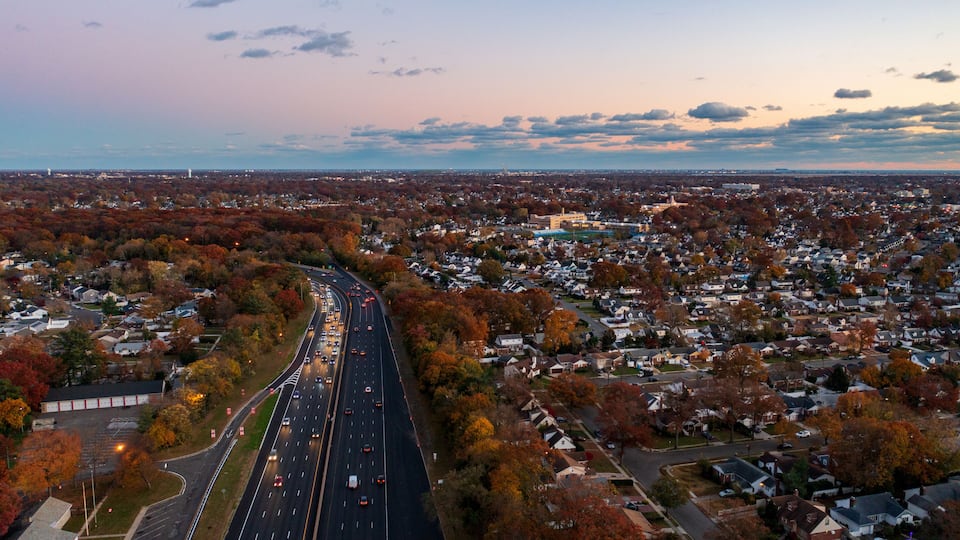 A highway curving through a suburban area with autumn-colored trees and a distant city skyline.