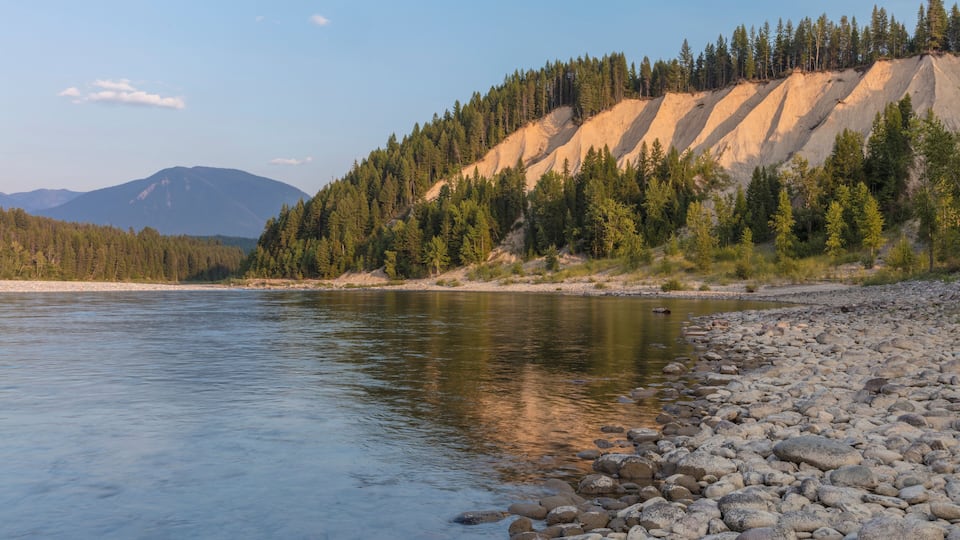 A river with a rocky shoreline, a forested hillside, and distant mountains.