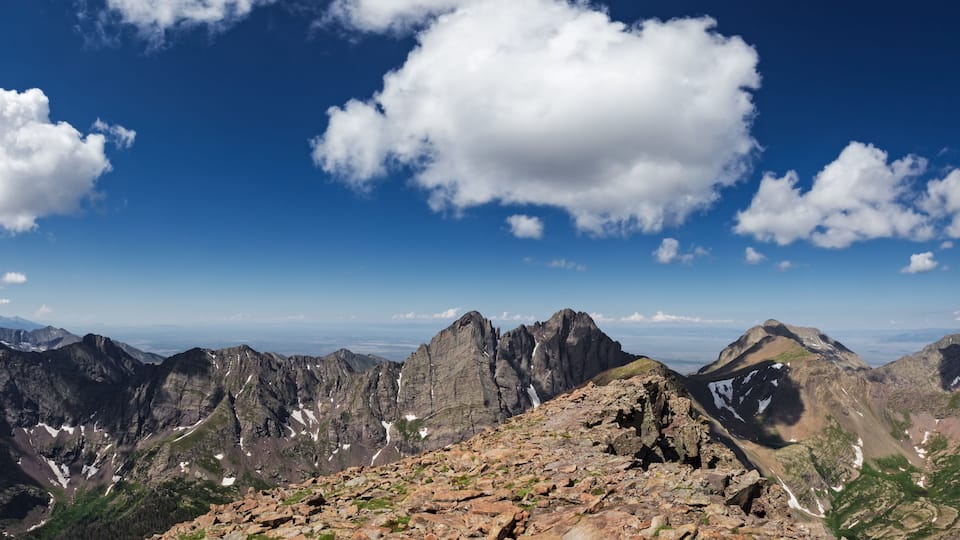 A mountain range with rocky terrain and snow patches under a clear blue sky with scattered clouds.