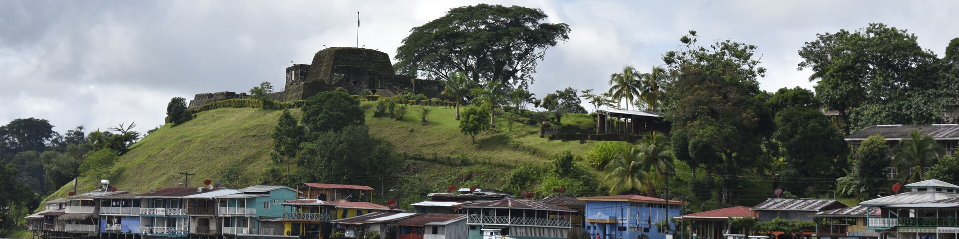 Uma vila às margens do rio, com casas coloridas, uma fortaleza no alto de uma colina e vegetação exuberante.