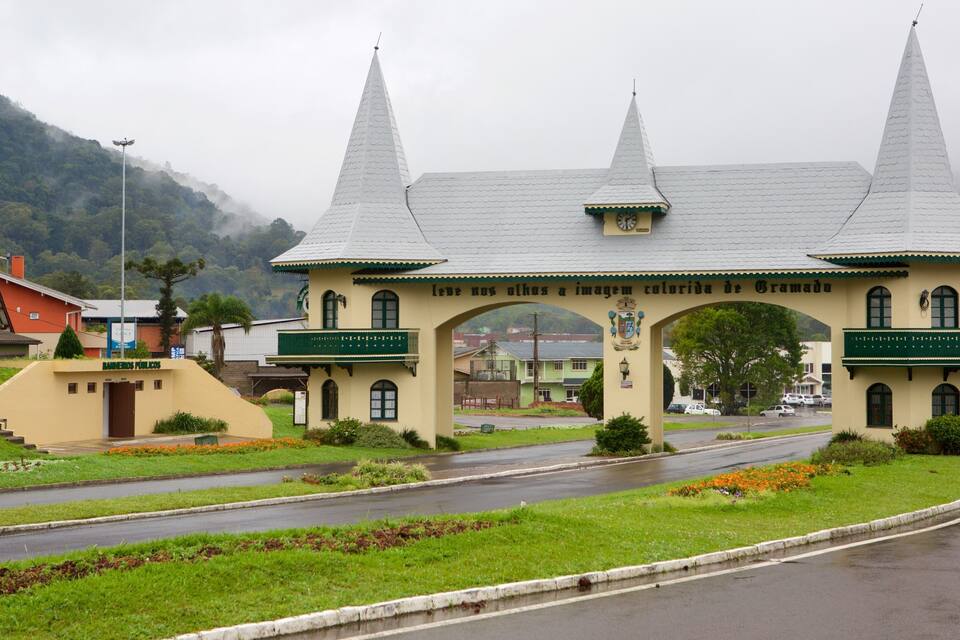 A historic building with a green archway and two spires, surrounded by lush greenery and a wet road.