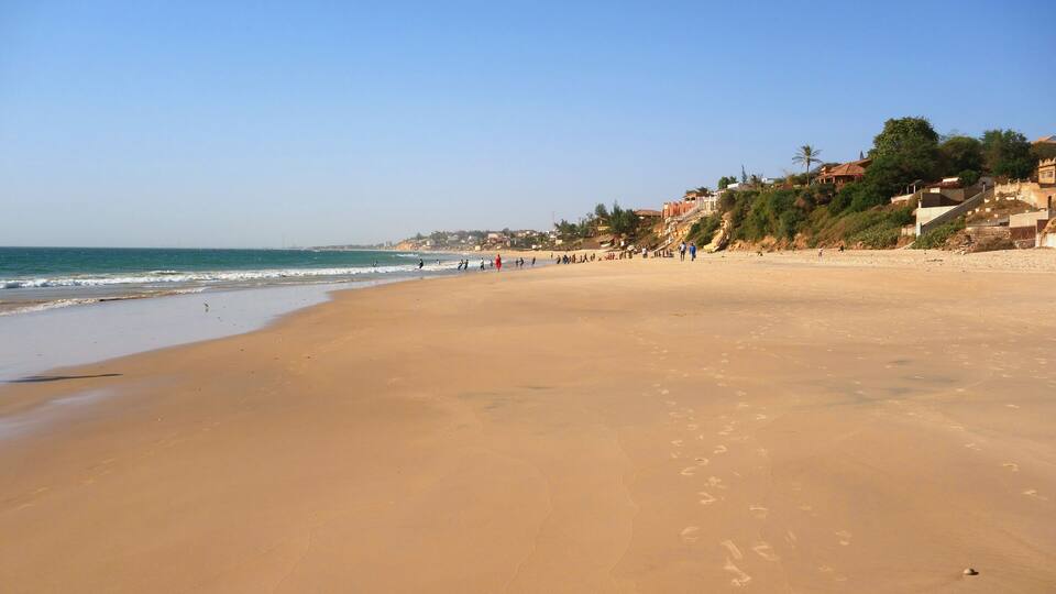 A wide sandy beach with footprints, a clear blue sky, and buildings on a hillside in the background.