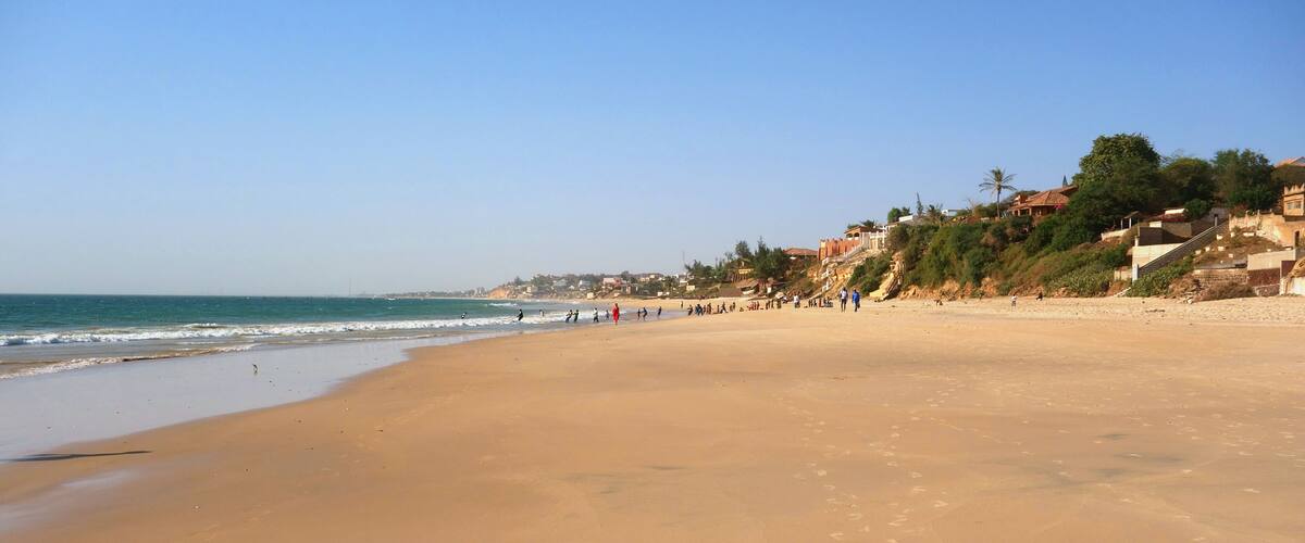 A wide sandy beach with footprints, a clear blue sky, and buildings on a hillside in the background.