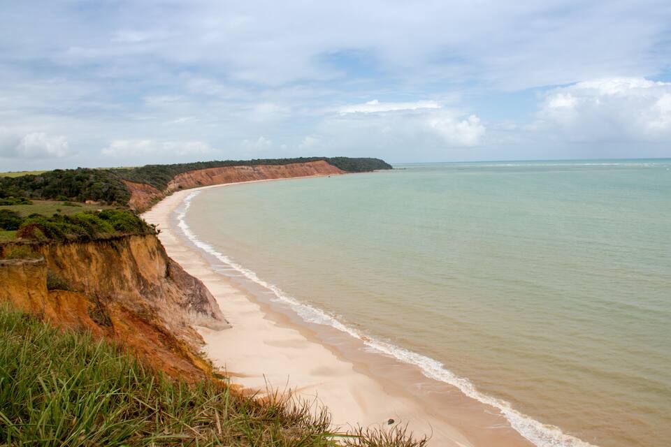 Paisagem costeira com uma praia arenosa, um penhasco com vegetação e um mar tranquilo sob um céu parcialmente nublado.