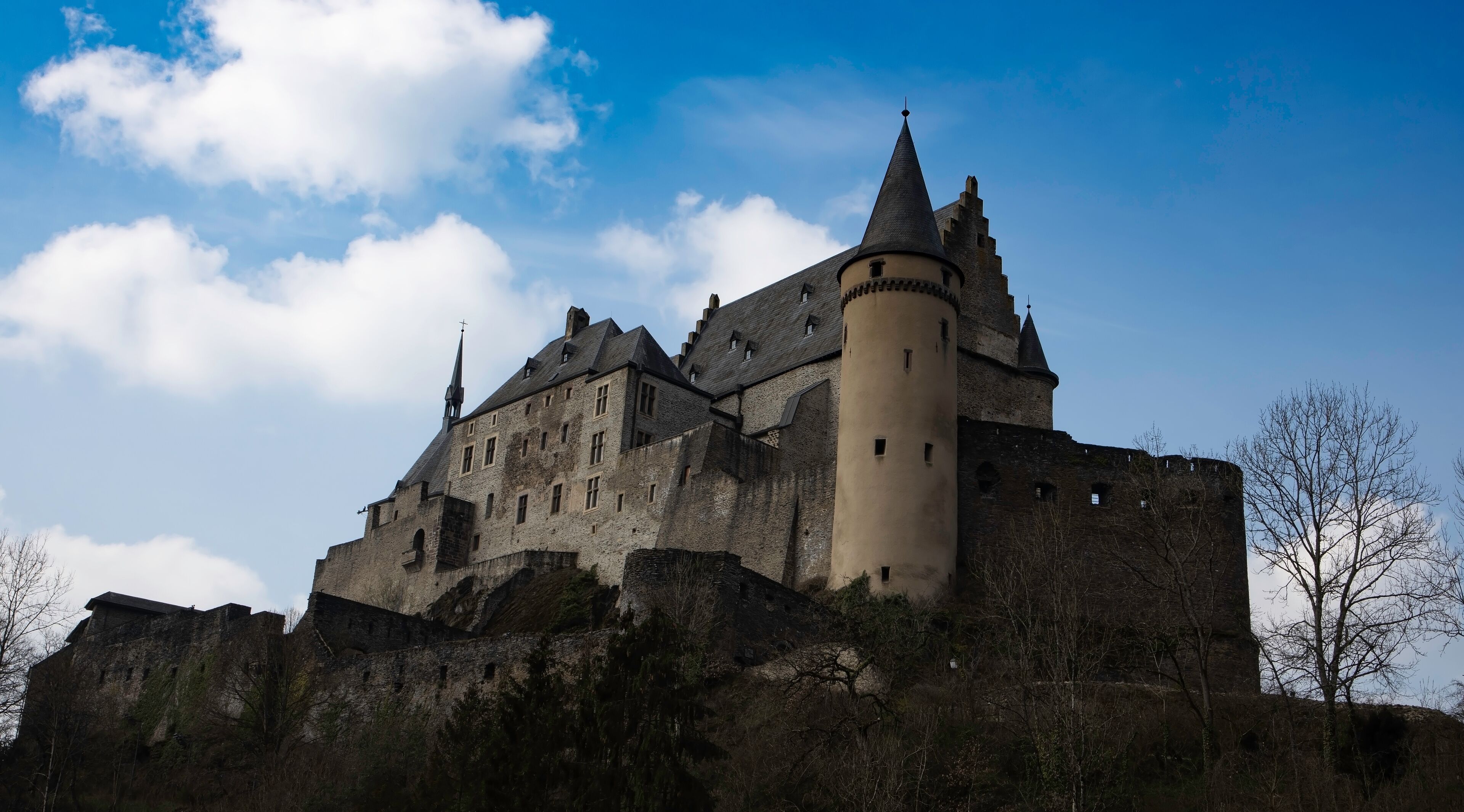 A historic castle with multiple towers and a prominent central tower, surrounded by a hilly landscape.