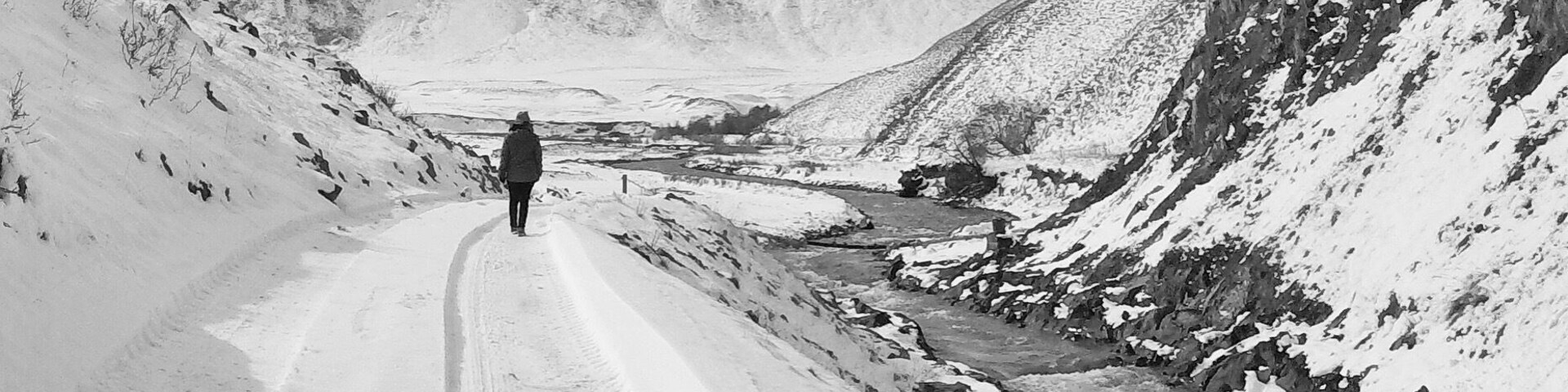 Uma estrada montanhosa coberta de neve, com uma pessoa caminhando se afastando da câmera.
