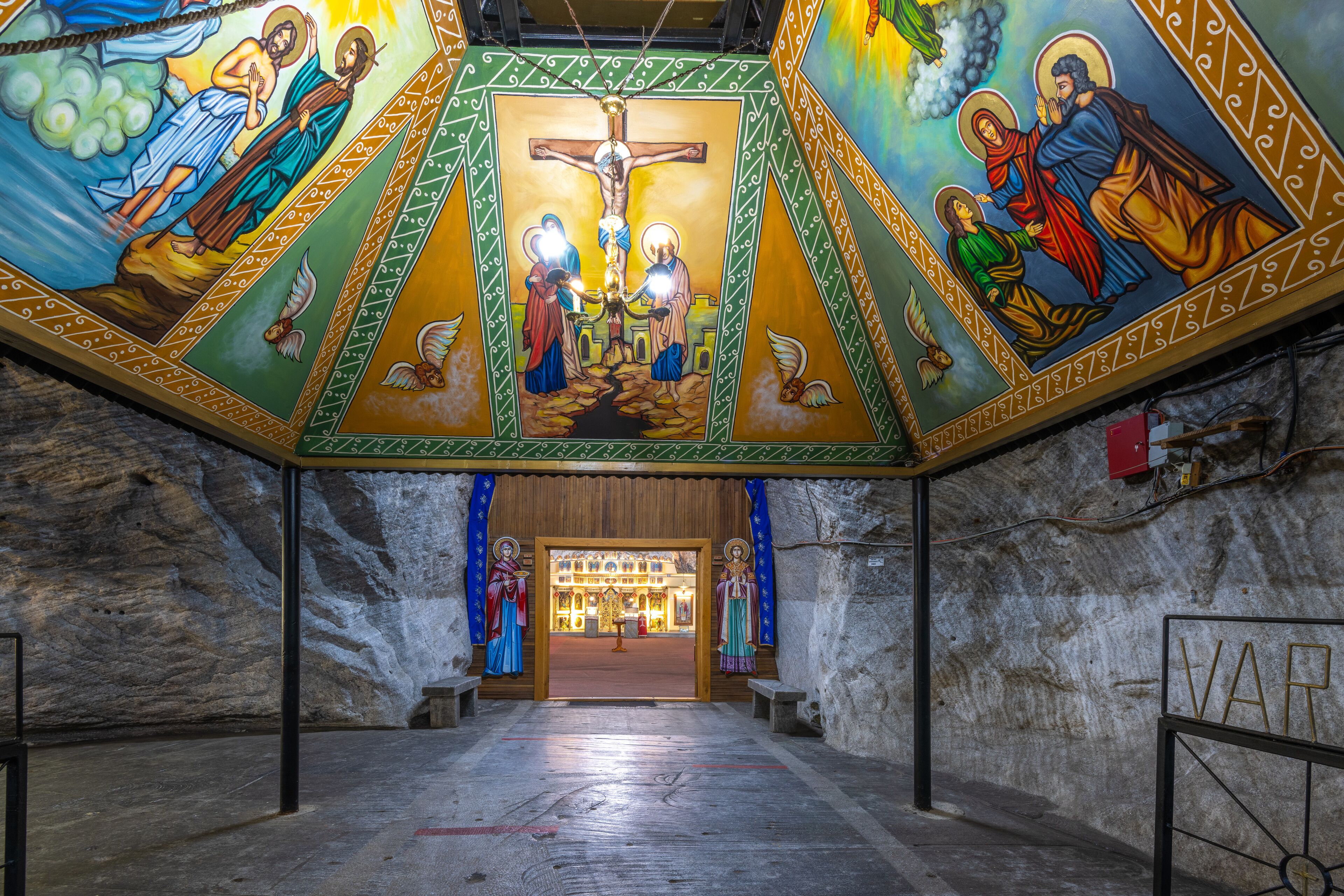 A church interior with a large crucifix, colorful frescoes, and stone walls.