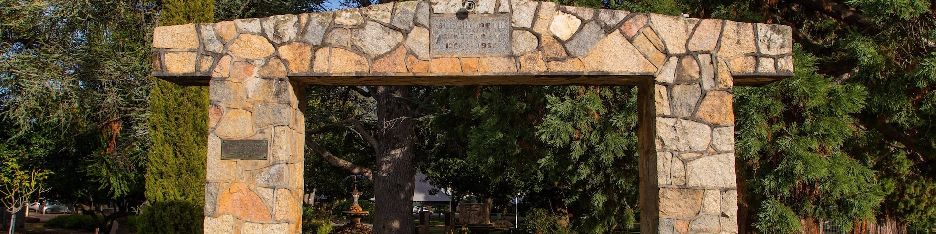 A stone archway with a plaque reading "FELDMOUTH CEMETERY 1854-1954" in a park with trees and a fountain.