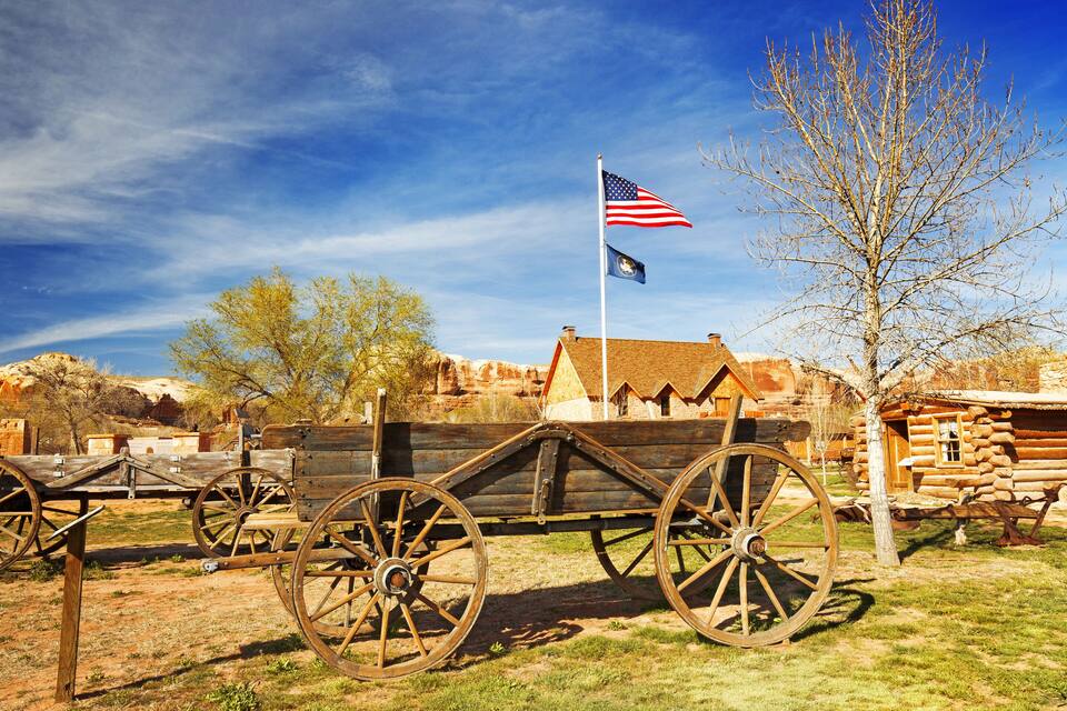 A wooden wagon with large wheels, an American flag, and a log cabin in a rural setting.