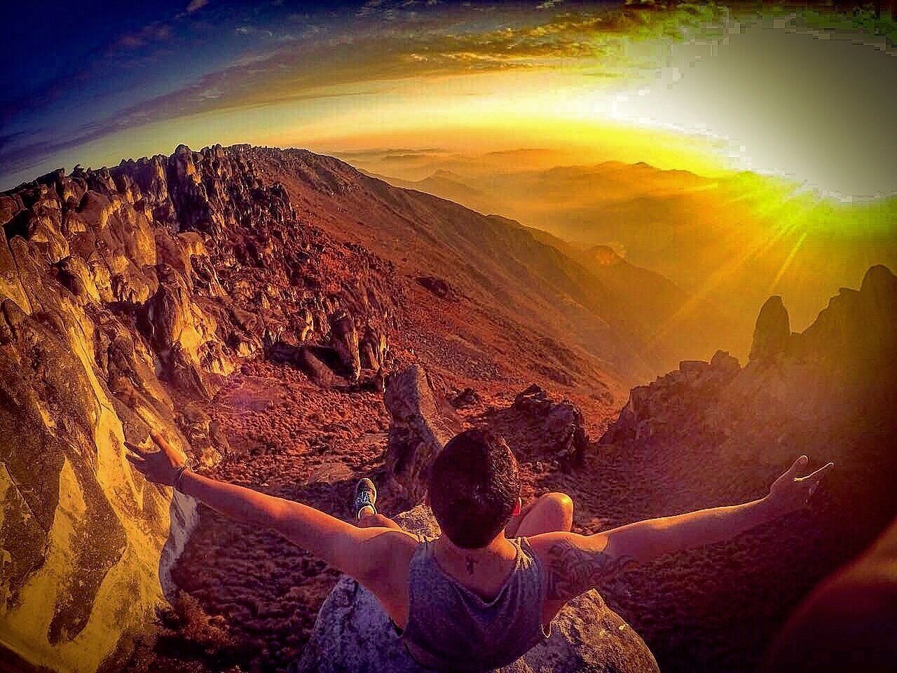 A person standing on a rocky mountain with arms outstretched, overlooking a vast mountainous landscape during sunset.