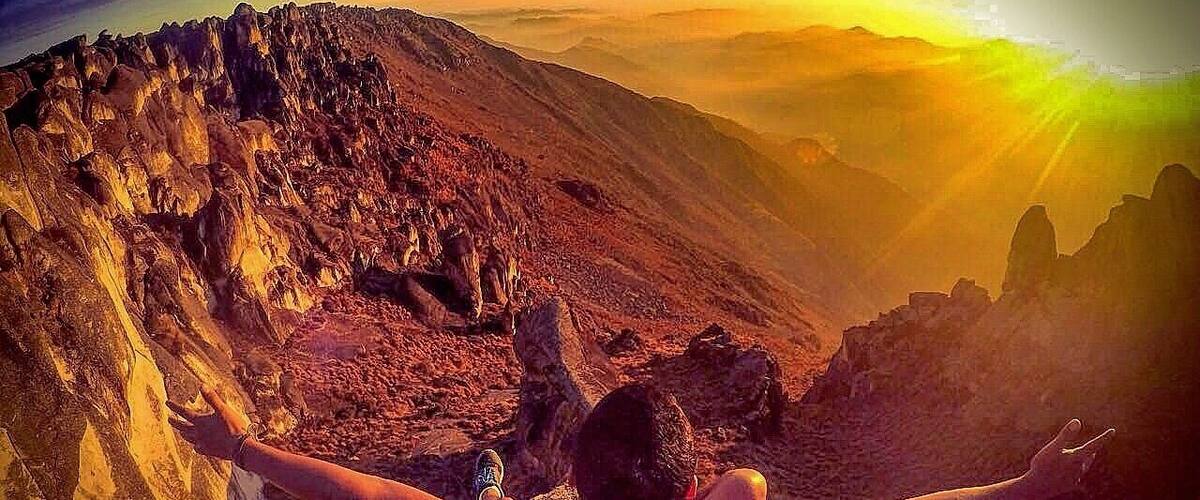 A person standing on a rocky mountain with arms outstretched, overlooking a vast mountainous landscape during sunset.
