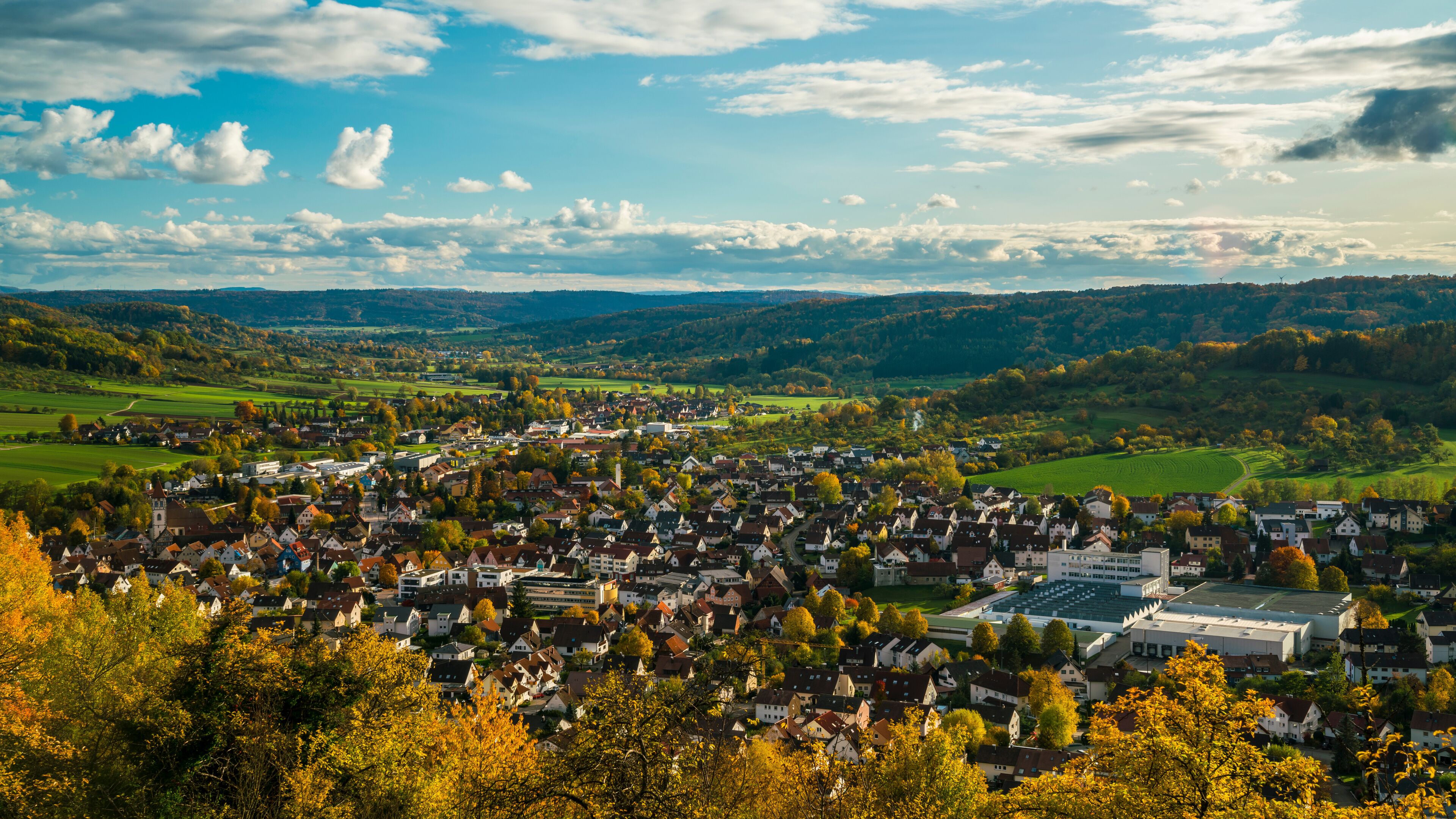 A town nestled in a valley with rolling hills in the background, surrounded by autumn-colored trees.