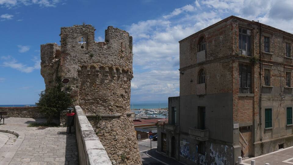 A historic stone building with a tower, a paved square with benches, and a view of the sea.