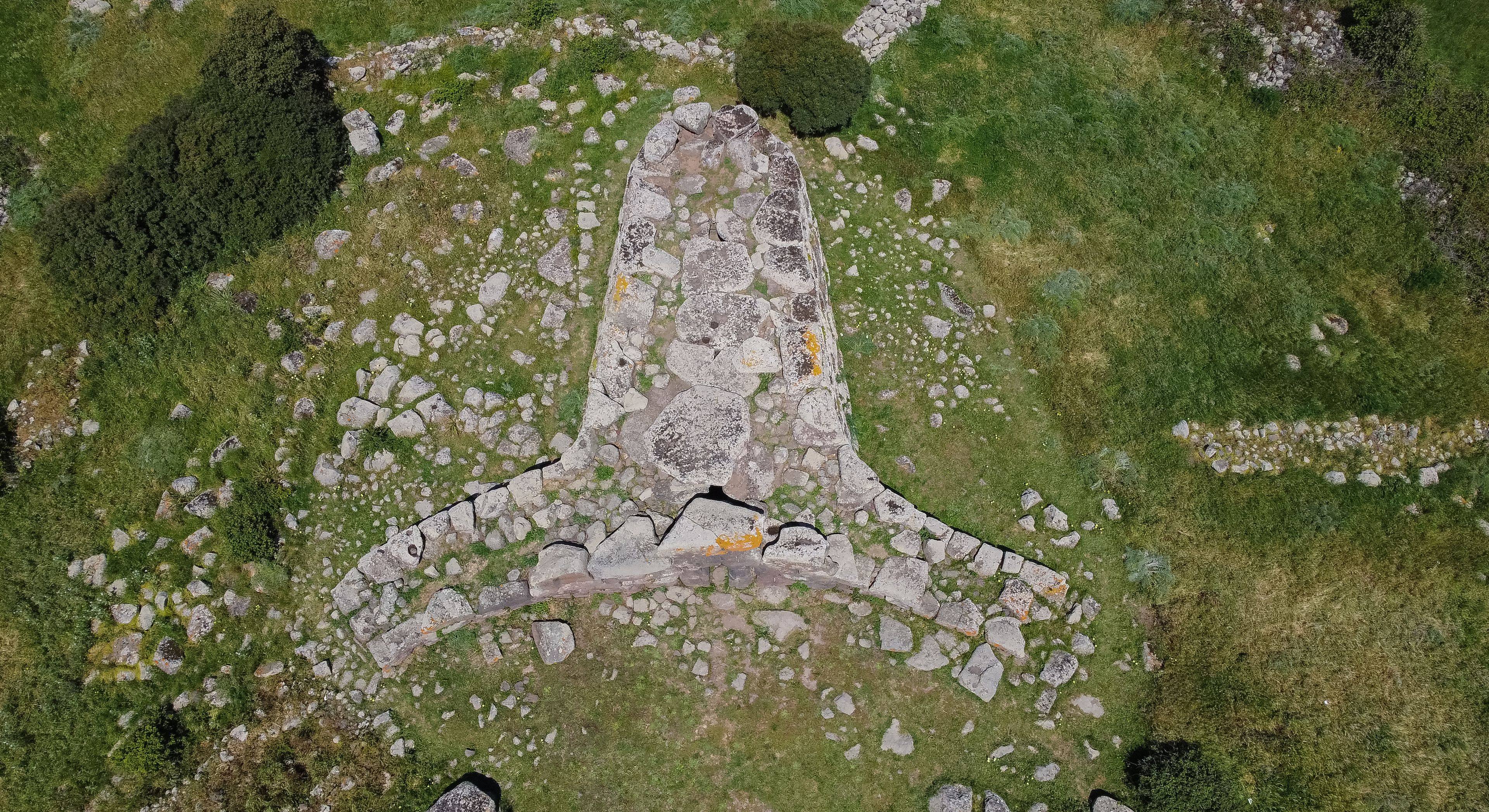 Vista aérea de uma construção de pedra com um círculo central de pedras, rodeado por pedras menores e grama.