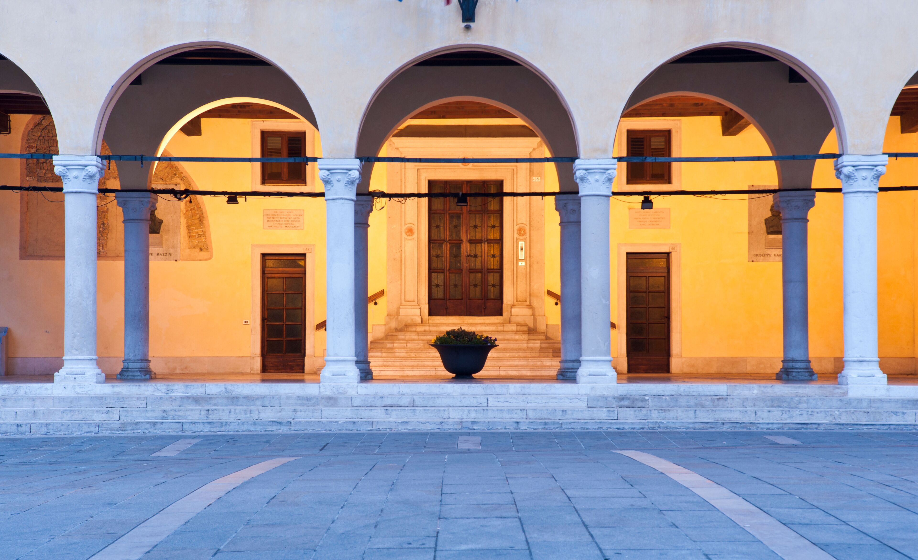 A building with three arched doorways, each with a wooden door and a potted plant in front.