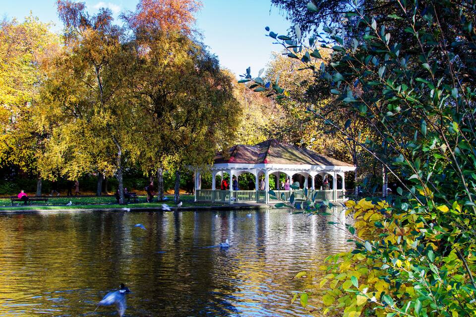 A gazebo by a lake with autumn foliage, ducks swimming, and people in the background.