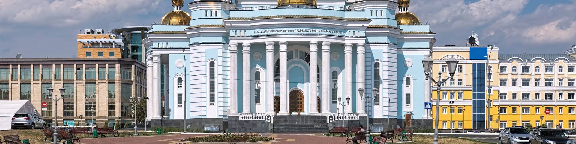 A large, ornate building with a golden dome and white columns, surrounded by a paved area with benches and flower beds.
