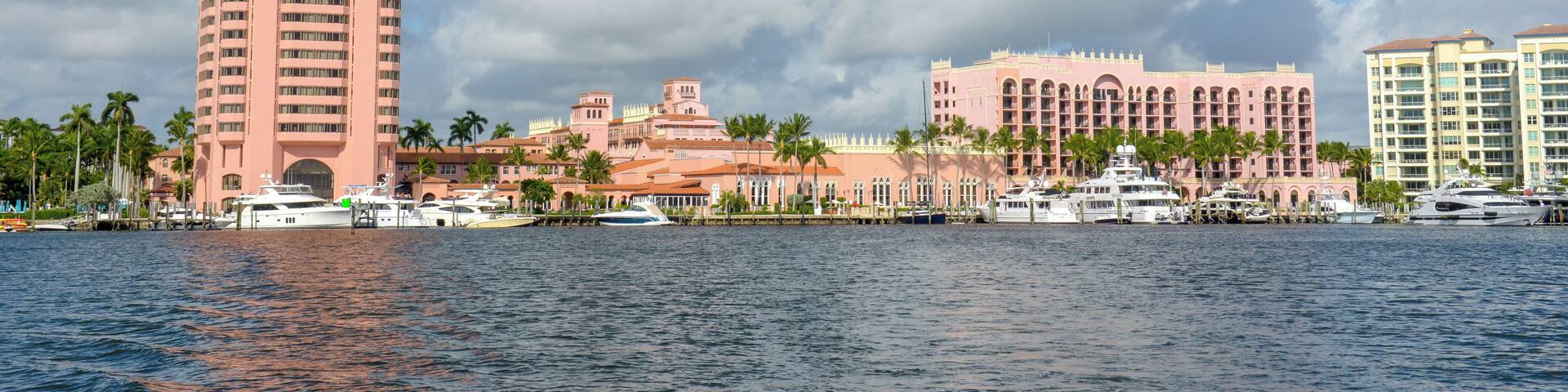 A waterfront with a tall pink building, several smaller pink and yellow buildings, and boats docked along the shore.