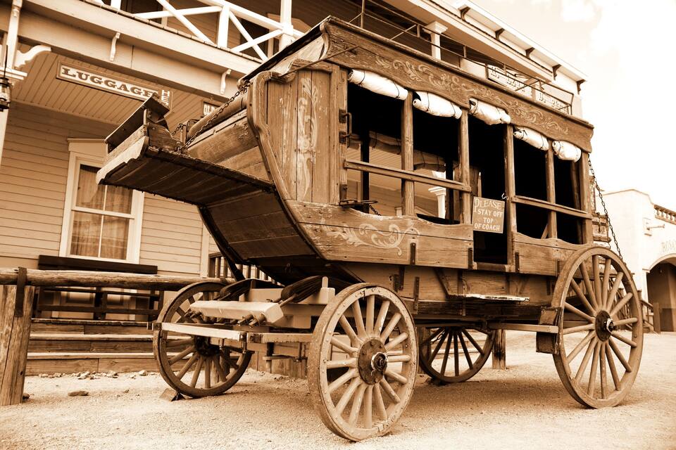 An old wooden carriage with large wheels parked in front of a building with a 'LUGGAGE' sign.