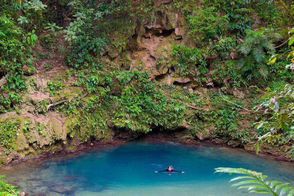A person kayaking in a clear blue river surrounded by lush greenery and rocky cliffs.