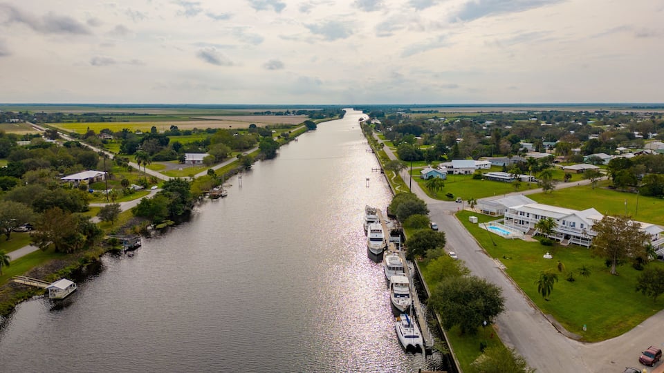 A river with boats docked on the right, surrounded by greenery and houses.
