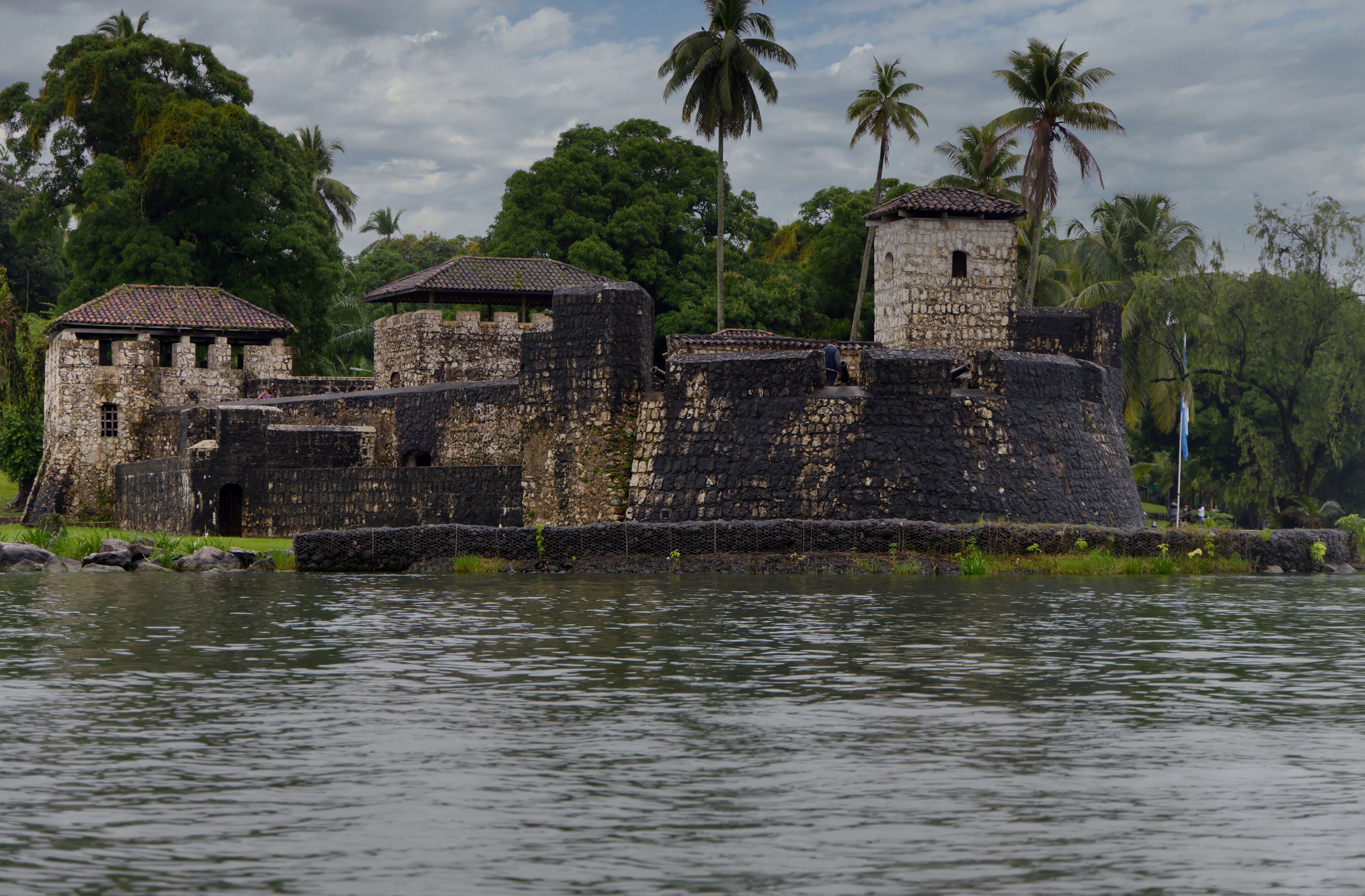 A historic stone fort with multiple towers and a surrounding moat, set against a backdrop of lush greenery and palm trees.