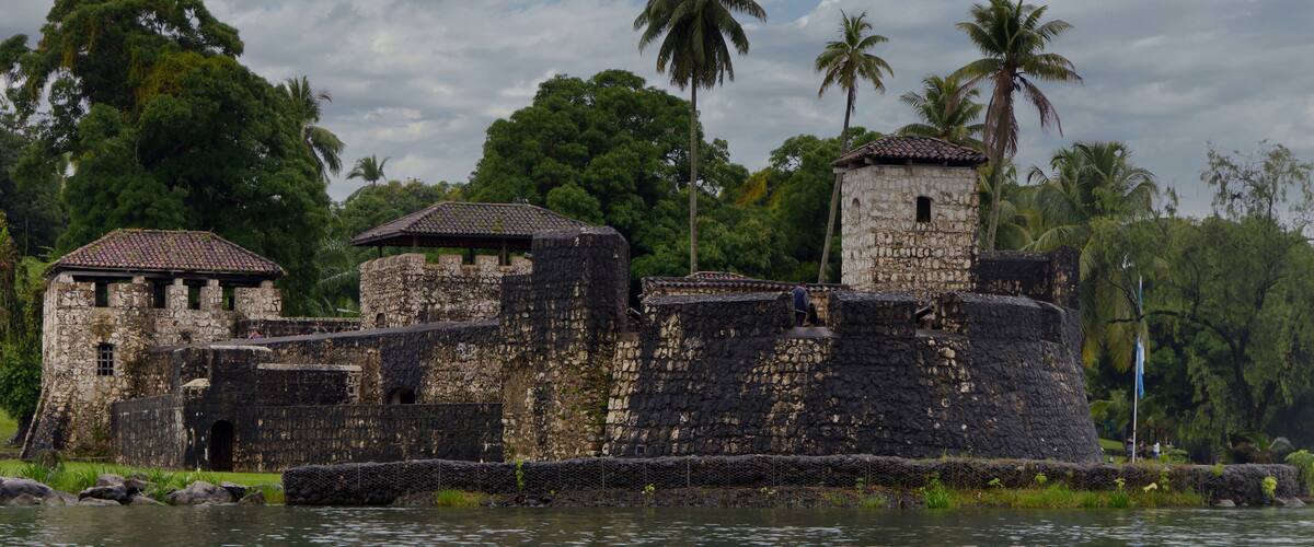 A historic stone fort with multiple towers and a surrounding moat, set against a backdrop of lush greenery and palm trees.