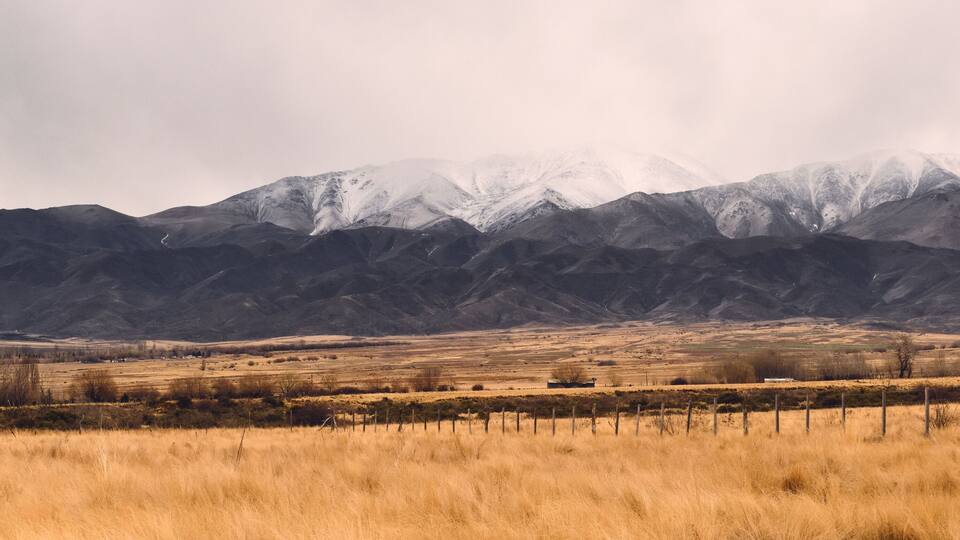 A mountainous landscape with snow-capped peaks, a dry grassy field, and a distant building.