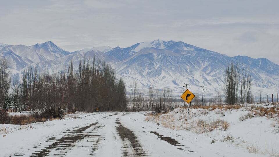 A snow-covered road with a mountain range in the background and a yellow warning sign.