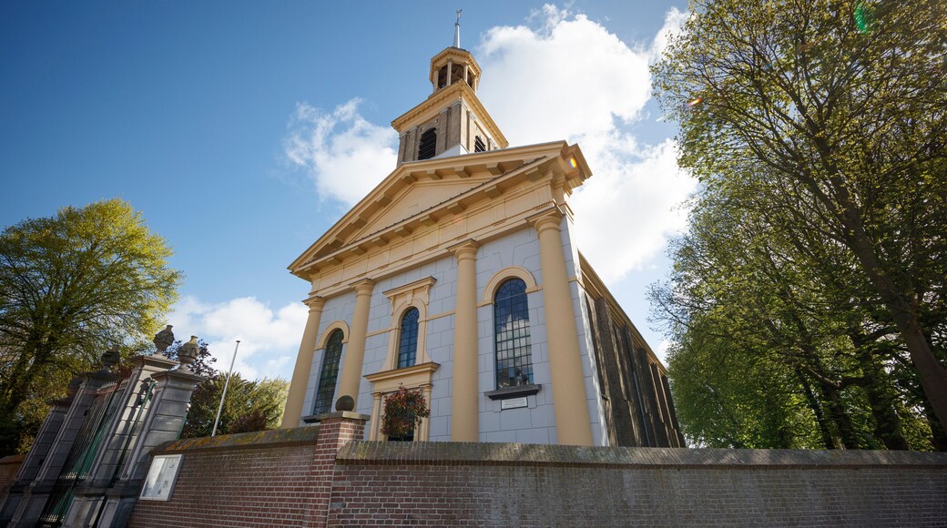 Low-angle shot of a light beige church with pale gray walls, tower, and decorative trim. Surrounding trees and a brick wall are visible. Sunny day.