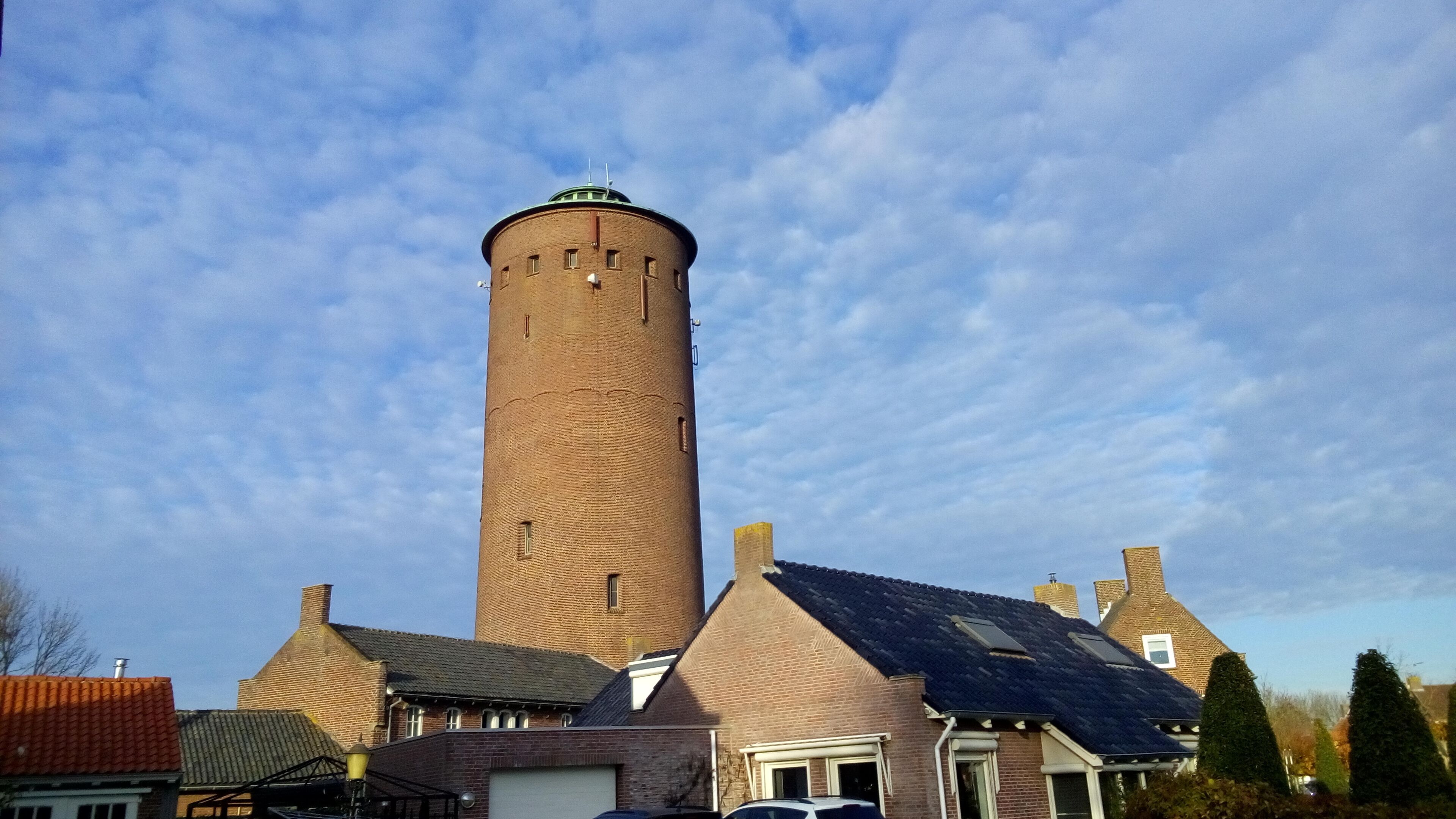 A tall brick tower with a conical roof, surrounded by houses with pitched roofs and a partly cloudy sky.