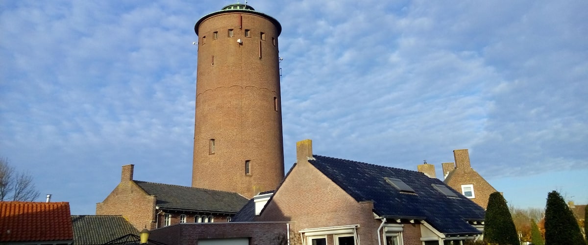 A tall brick tower with a conical roof, surrounded by houses with pitched roofs and a partly cloudy sky.
