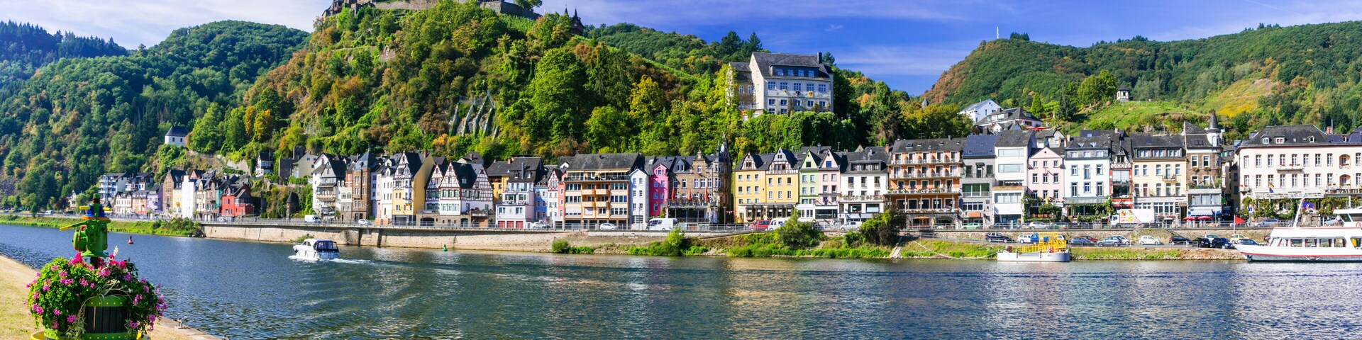 Uma cidade ribeirinha com edifícios coloridos, um castelo no topo de uma colina e barcos na água.