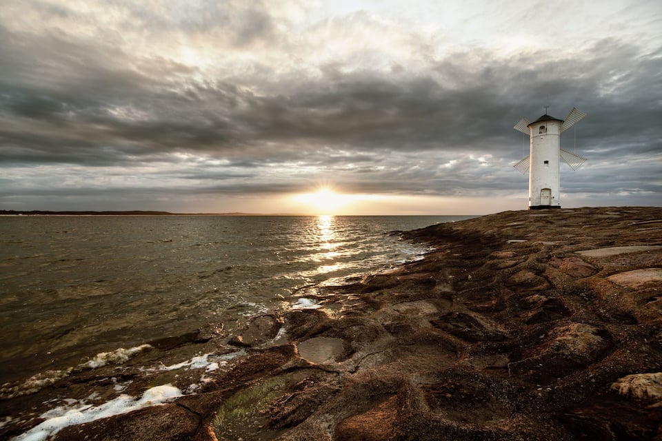 Un phare se dresse sur une côte rocheuse, avec le soleil couchant à l’horizon.