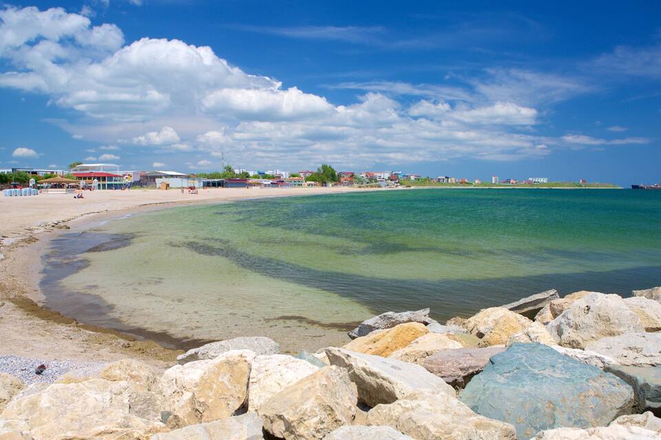 A beach with clear water, rocks in the foreground, and buildings along the shore.