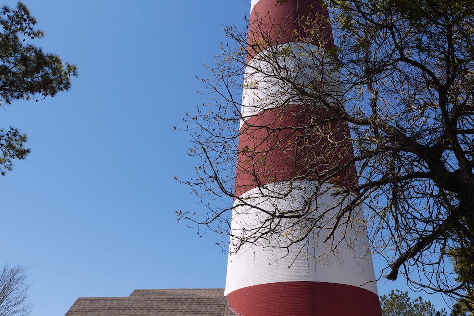 A tall red and white lighthouse with a black top, surrounded by trees and a small building.