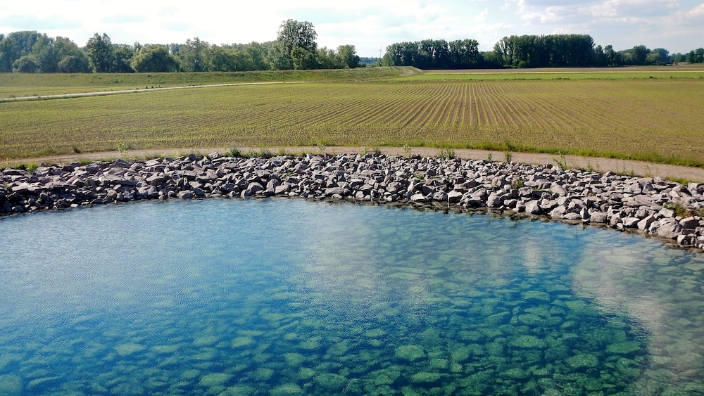 A clear pond surrounded by a rocky edge, with fields and trees in the background.