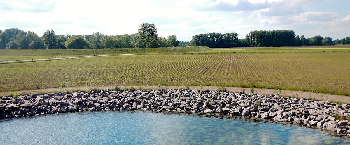 A clear pond surrounded by a rocky edge, with fields and trees in the background.