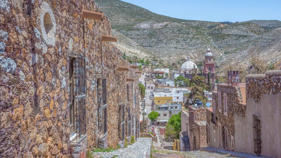 A narrow stone street flanked by buildings with stone walls and a church with a dome in the distance.