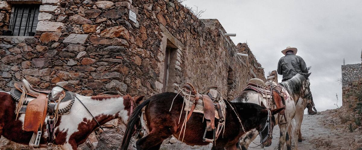 A stone building with a sign, a person on horseback, and two horses with saddles.