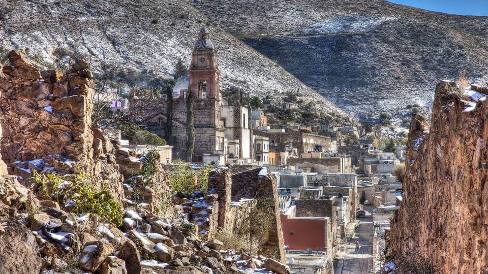 A mountainous town with a prominent church tower, rocky terrain, and snow patches.