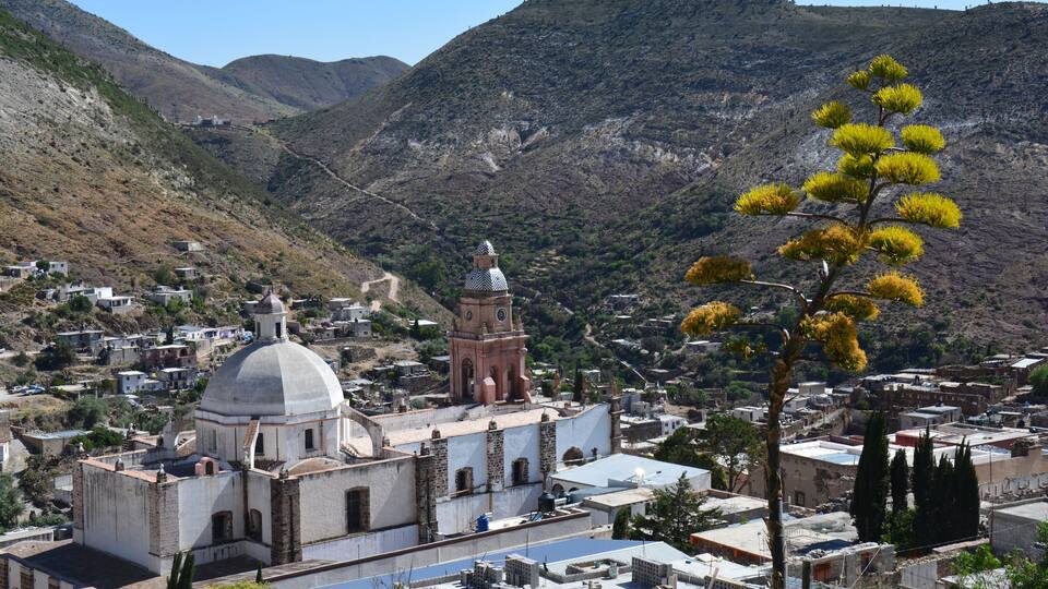 A town with a prominent church featuring a dome and bell tower, surrounded by mountainous terrain and sparse vegetation.