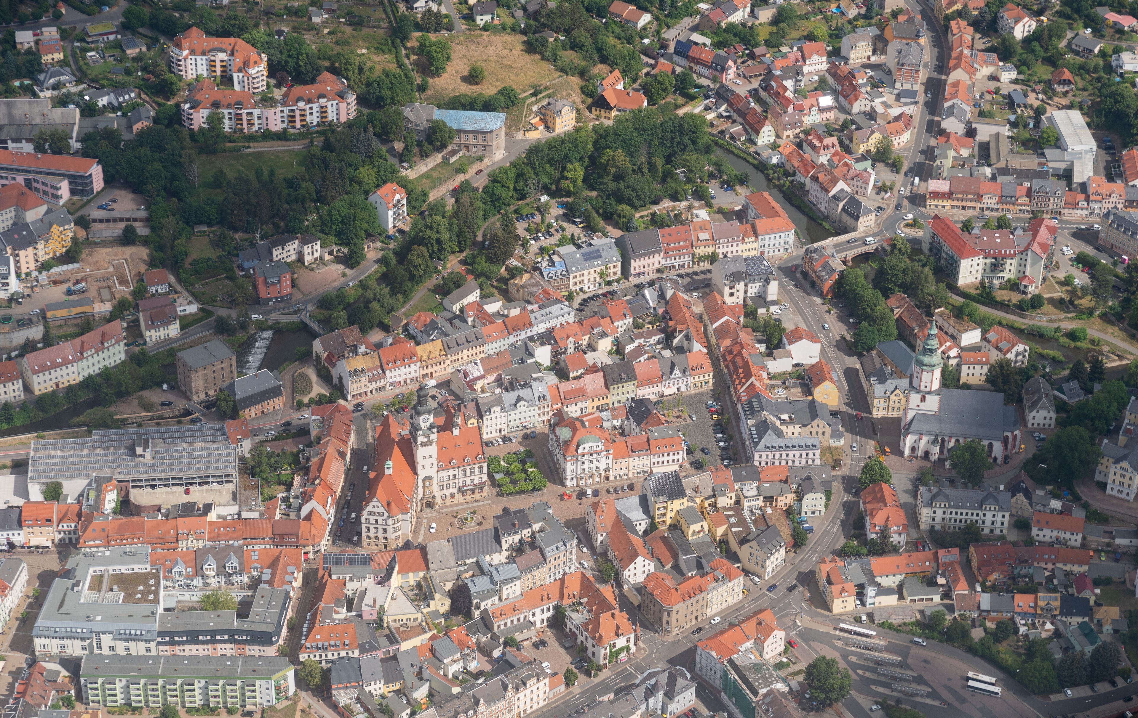 An aerial view of a densely built town with red-roofed buildings, a river, and a church with a clock tower.