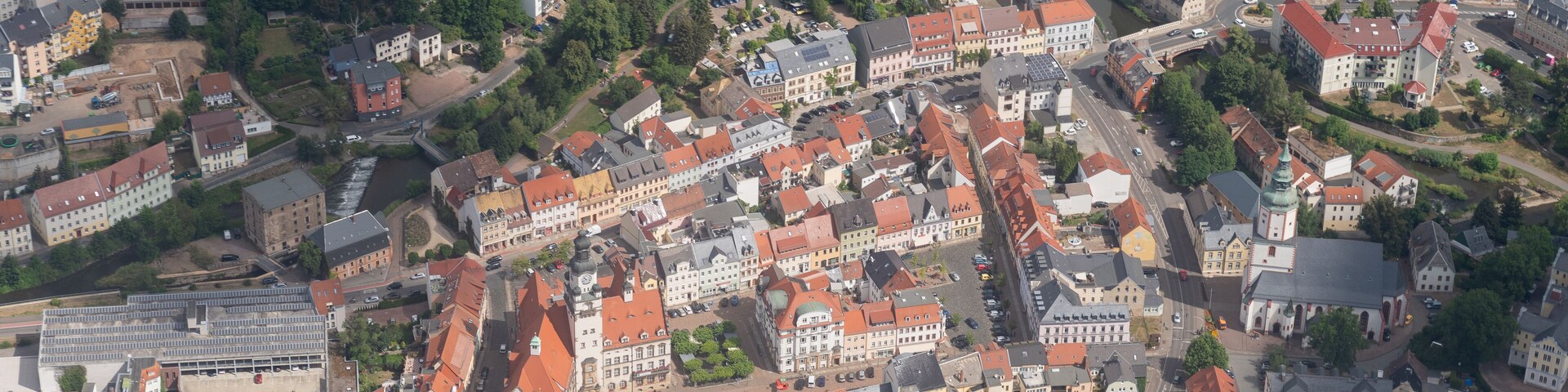An aerial view of a densely built town with red-roofed buildings, a river, and a church with a clock tower.