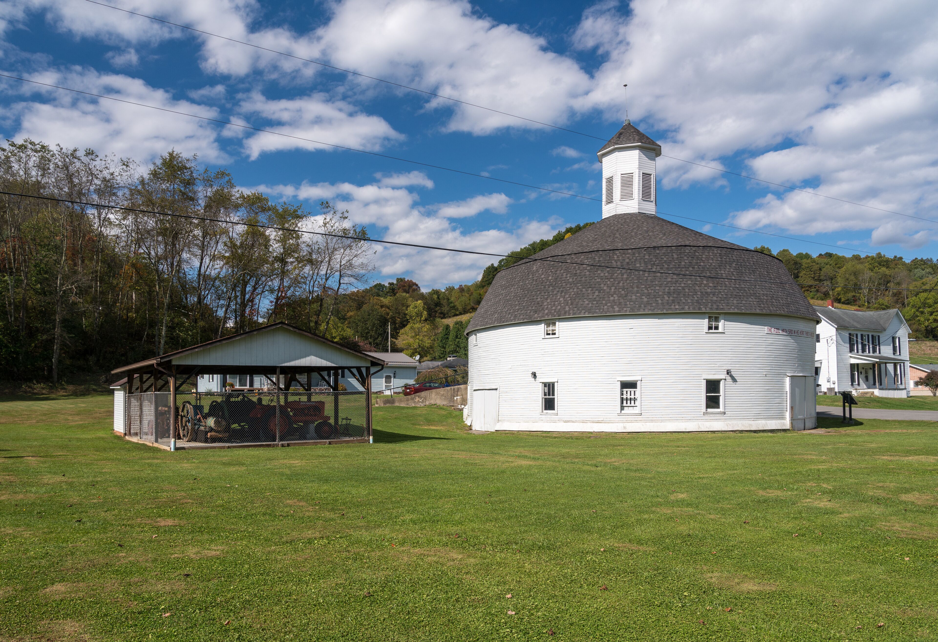 A white historic building with a conical roof and a small tower, surrounded by a grassy field and trees.