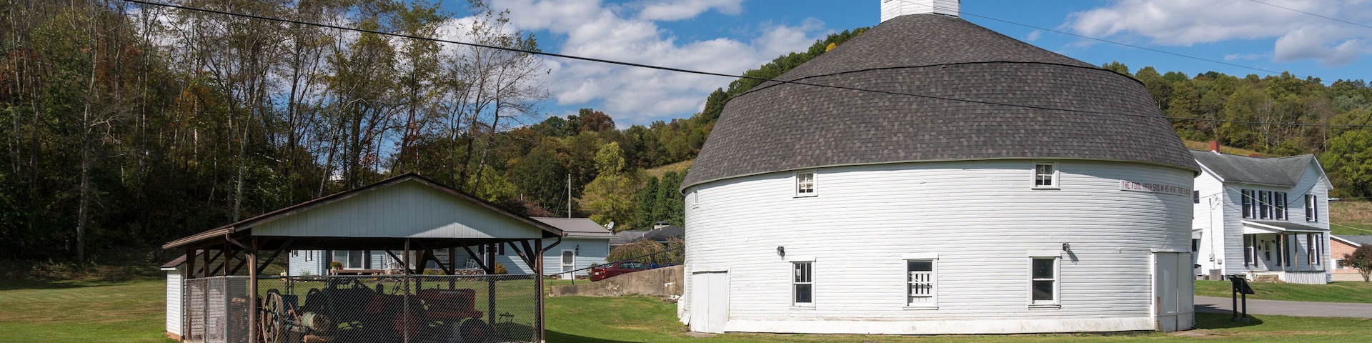 A white historic building with a conical roof and a small tower, surrounded by a grassy field and trees.
