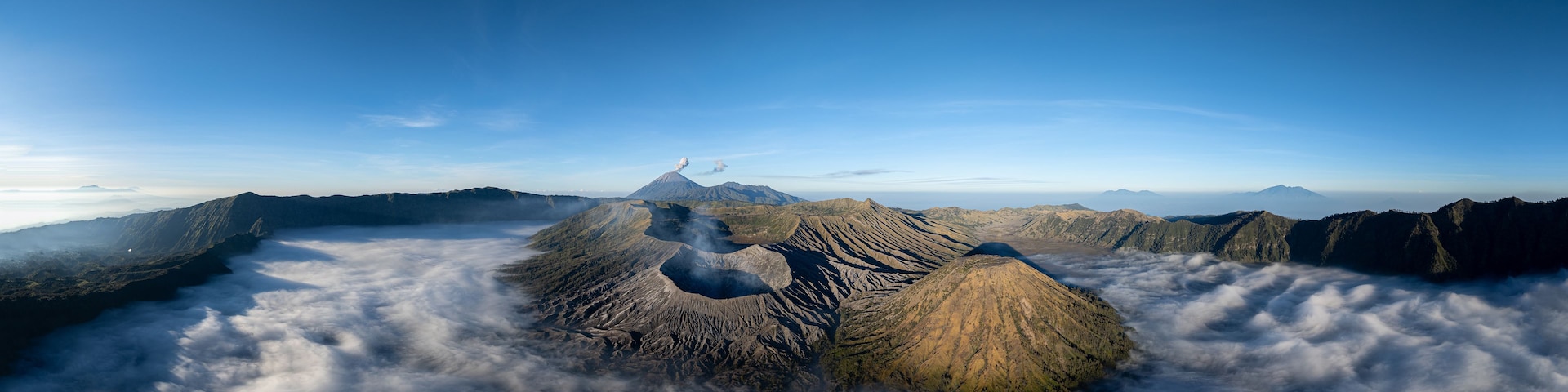 Una cordillera con un pico destacado que emite humo, rodeada de nubes y montañas lejanas bajo un cielo despejado.