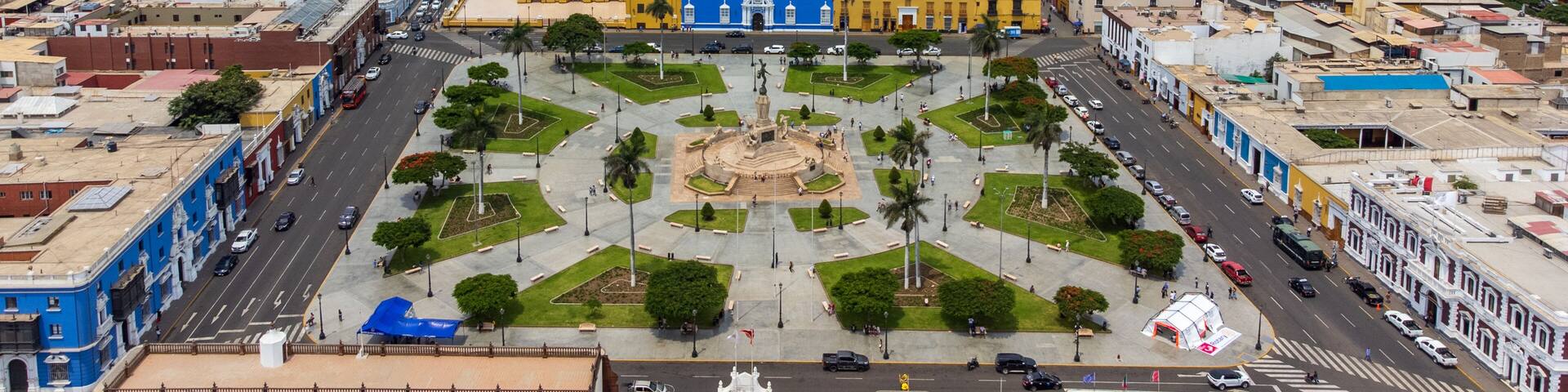 Un paisaje urbano con una catedral amarilla destacada, una plaza central con una fuente y montañas al fondo.